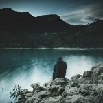 a man sits on a rock staring across a lake at dusk