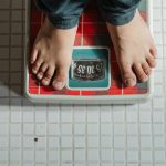 From above crop anonymous barefoot child in jeans standing on weigh scales on tiled floor of bathroom
