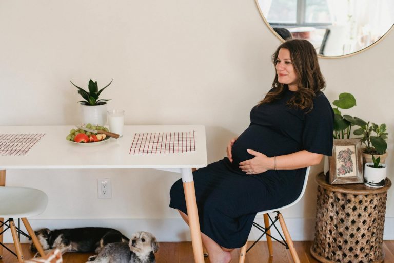 Side view of positive pregnant female in casual clothes touching belly and sitting at table while chilling at home with dogs in daytime
