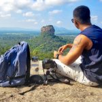 Man Sitting on Top of Gray Cliff Mountain Beside Backpack, Water Bottle, and Camera