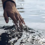 A serene close-up of a hand gently touching a sparkling water surface under daylight.