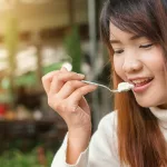Happy woman enjoying a creamy dessert in a sunlit outdoor setting.